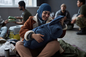 Front view portrait of young Middle Eastern woman playing with baby sitting on floor at refugee shelter, copy space
