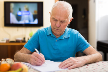 Focused senior man reading and signing papers at home table