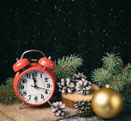 Red retro alarm clock with fir branches and a Christmas tree toy. The clock shows a few minutes until New Year.