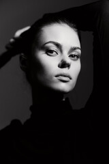 Portrait of a confident young woman with a sleek hairstyle in a black turtleneck, striking a pose against a dark background. Emphasizing strength and elegance.