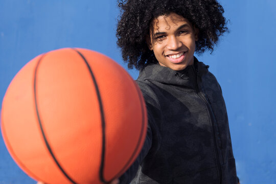 positive teen african american male with curly hair offering hand with basketball ball towards camera against blue background, concept of sport and young lifestyle
