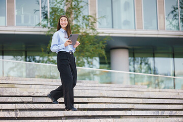 A Young Woman Walks Down Outdoor Stairs Holding a Tablet While Smiling in a Modern Urban Setting During Daytime