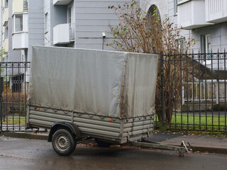Large two-wheeled gray awning trailer for a passenger car parked at the fence of an apartment building