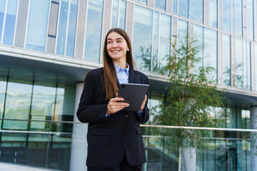 Fototapeta premium Professional Woman in Business Attire Smiles While Holding a Tablet Outside a Modern Office Building