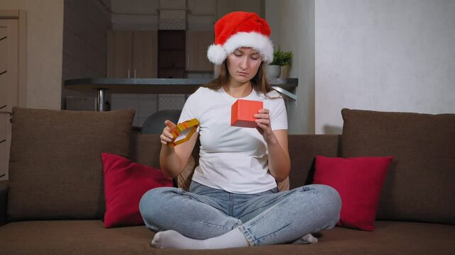 Woman in Santa hat sitting on sofa opening christmas gift. Frustration washes over her face as she opens contents inside, leading to moment of sadness, she is dissatisfied.
