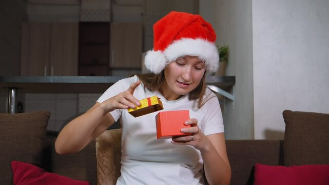 Woman in Santa hat sitting on sofa opening christmas gift. Frustration washes over her face as she opens contents inside, leading to moment of sadness, she is dissatisfied.