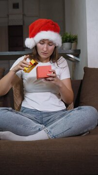 Woman in Santa hat sitting on sofa opening christmas gift. Frustration washes over her face as she opens contents inside, leading to moment of sadness, she is dissatisfied. vertical video