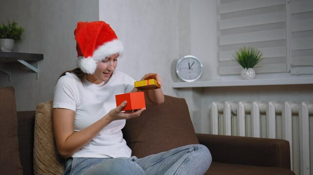 Woman in Santa hat sitting on sofa opening christmas gift. Frustration washes over her face as she opens contents inside, leading to moment of sadness, she is dissatisfied.