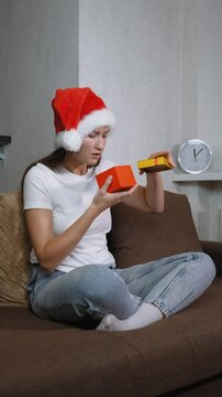 Woman in Santa hat sitting on sofa opening christmas gift. Frustration washes over her face as she opens contents inside, leading to moment of sadness, she is dissatisfied. vertical video
