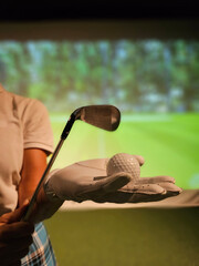 Golfer in glove holds golf ball and club indoors. Playing golf on a simulator vertical concept