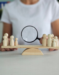 Person holding a magnifying glass over wooden figurines on a balance scale representing focus,...
