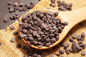 Choco chips for melting as an ingredient for baking. Chocolate bits in wooden spoon with rough burlap on a brown board
