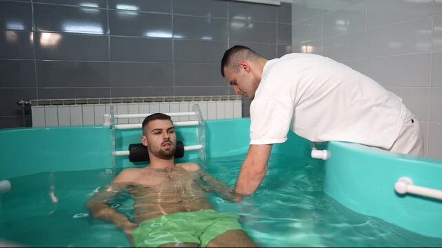 Young man receiving hydrotherapy treatment in a spa facility, with a professional therapist gently exercising his arm in a therapeutic tub. Concept of rehabilitation and physical therapy