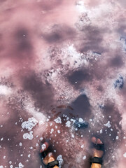 Feet in pink saltwater lake with foam and clouds reflection