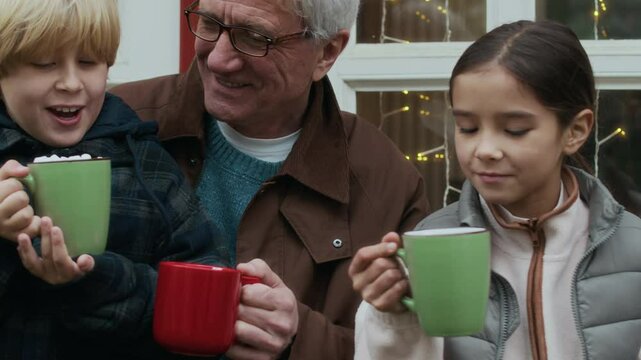 Chest up of senior man in glasses with two young grandchildren holding mugs with hot chocolate and marshmallows sitting and talking on cozy porch outside of red house, decorated with string lights