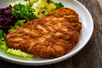 Crispy breaded fried pork chop, boiled potatoes and red cabbage served on white plate on wooden table
