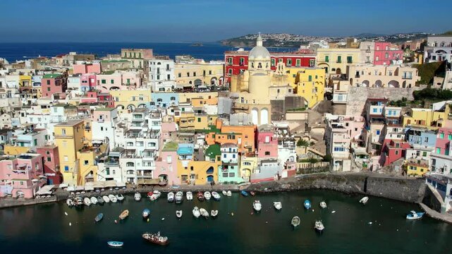 Breathtaking Procida Corricella. Aerial 4K drone flyover from yellow church along colorful pastel houses to hilltop castle. Gulf of Naples Italy travel scenery. iconic summer Mediterranean coast