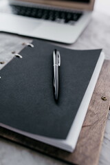 Handmade notebook in wooden cover with black watercolor paper and black metal pen near laptop on grey textured table