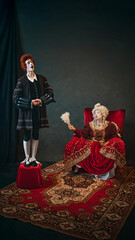 Young boy, court servant in red wig standing next to elderly woman, medieval empress and singing to her, set against dark vintage background. Concept of history, comparison of eras