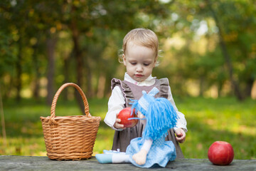 Little girl with basket, doll and apples playing in autumn park