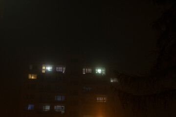 Naklejka premium On a mysterious foggy night, the soft glow of illuminated apartment windows emerges, casting a haunting yet captivating ambiance throughout the dark cityscape, creating an eerie yet serene atmosphere
