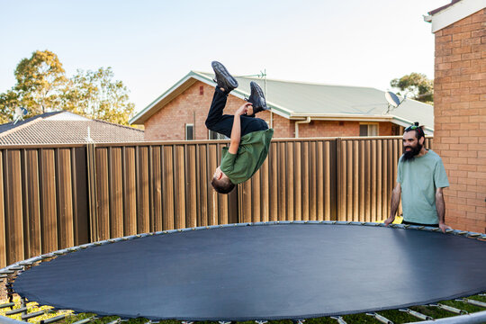 Dad watching eight year old Aussie kid jumping and doing flips on trampoline in backyard