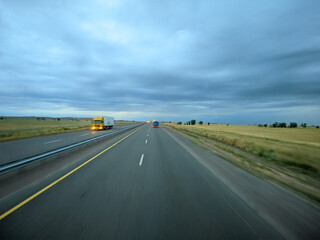 Evening road with cars and trucks. Photo in motion. Cars drive fast and illuminate the road with their headlights. View of summer or autumn landcape with road on the field.