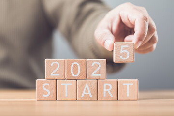 Businessman arranges wooden blocks spelling out 'START 2025' on a table, symbolizing the beginning...