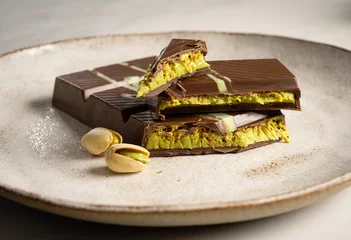 A broken bar of Dubai chocolate lies appetizingly on a ceramic plate and the delicious pistachio cream between the chocolate is clearly visible. © Anna
