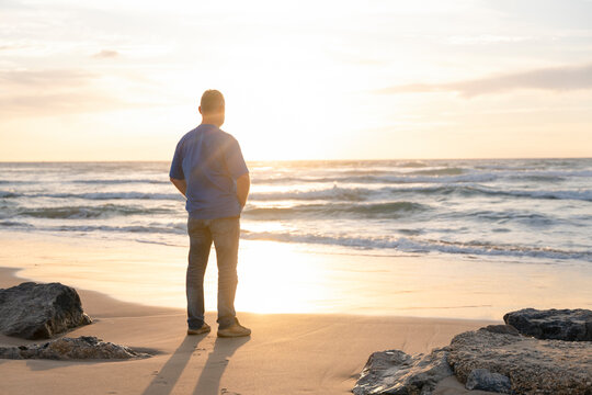 A man gazes at the sunset over the ocean while standing on the beach during a calm evening in nature