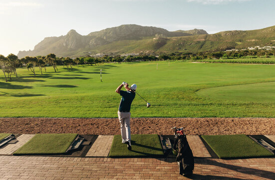 Man hitting a golf ball at the driving range of a golf club with mountain backdrop on a sunny day