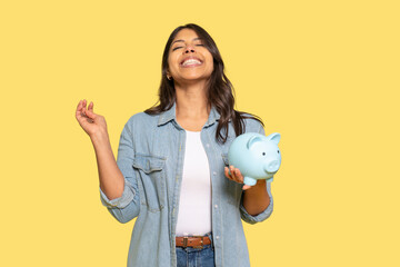 Happy woman holding a piggy bank against a bright yellow background while celebrating financial...
