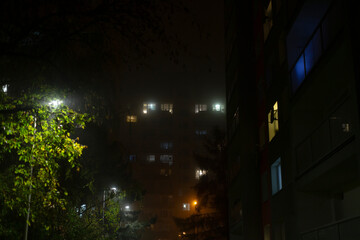 On a foggy night, the scene captivates with beautiful illuminated windows glowing brightly from the apartment buildings, surrounded by the soft glow of streetlights and lush green foliage all around