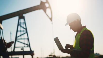 oil business. a worker works next to an oil pump holding a laptop. industry business oil and gas concept. lifestyle engineer studying the level of oil production on a laptop silhouette at sunset