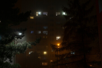 On a mysterious and foggy night, an apartment building stands prominently, softly illuminated by warm, inviting lights, creating a serene and peaceful atmosphere within the bustling urban environment