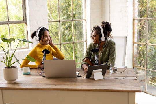 Two multiracial female friends recording podcast at home, smiling and wearing headphones
