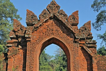 Naklejka premium Ancient red brick archway surrounded by lush greenery under a clear blue sky.