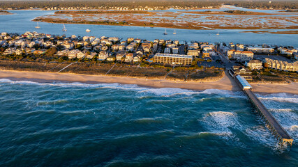 Aerial view of a pier stretching into the ocean at Wrightsville Beach, NC, surrounded by beachfront residential buildings and an inlet in the background.