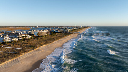 Aerial view of Wrightsville Beach, NC, featuring a long, sandy beach, ocean waves, a pier extending into the water, and nearby coastal homes and buildings.