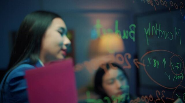 Two young businesswomen brainstorm ideas using sticky notes and markers on a glass wall in their office at night, showcasing teamwork and collaboration in a modern workspace