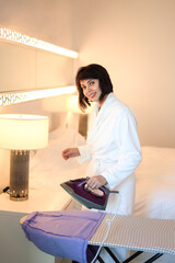 adult woman in a white robe irons her clothes in the bedroom.