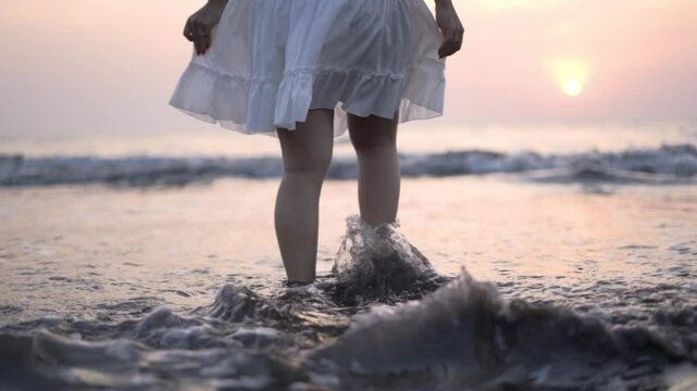 young woman slowly walking in shallow sea water on the beach wearing a short white dress with waves touching her feet and legs in slow motion