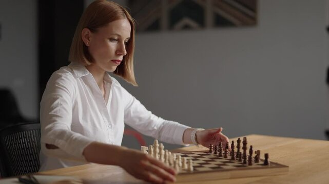 A focused business woman in a white shirt contemplates her next move while playing chess alone. She sits at her desk, strategizing her next move. 