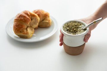 A woman's hand holding yerba mate tea with hot water, a traditional South America infusion. Beside her is a typical argentinian croissants called medialuna, a traditional breakfast in Argentina	