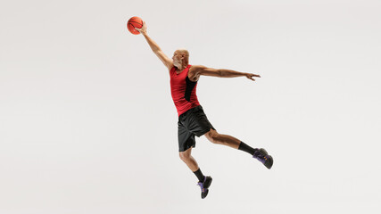 Young man in athletic clothing, wearing tank top and shorts, jumps with basketball toward hoop against grey studio background. Concept of sport event, competition, active and healthy lifestyle, game.