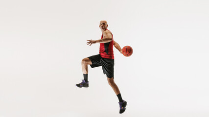 Athletic man in sports gear leaps with basketball in hand, preparing to make basket against grey studio background. Concept of sport event, competition, active and healthy lifestyle, game.