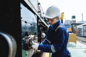 A man in a blue and white uniform is working on a train engine. He is wearing a white helmet and...