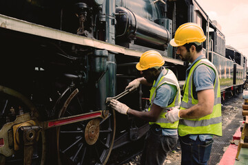 Two men in yellow helmets are working on a train engine. One of them is holding a wrench