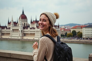 Fototapeta premium Smiling young woman in warm clothing exploring budapest with iconic parliament building in background.