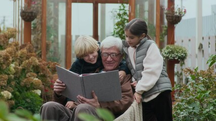 Medium shot of happy grandfather and his two young grandkids reminiscing cherishing moments by looking at photos in family album outdoors in lush garden with greenhouse - Powered by Adobe